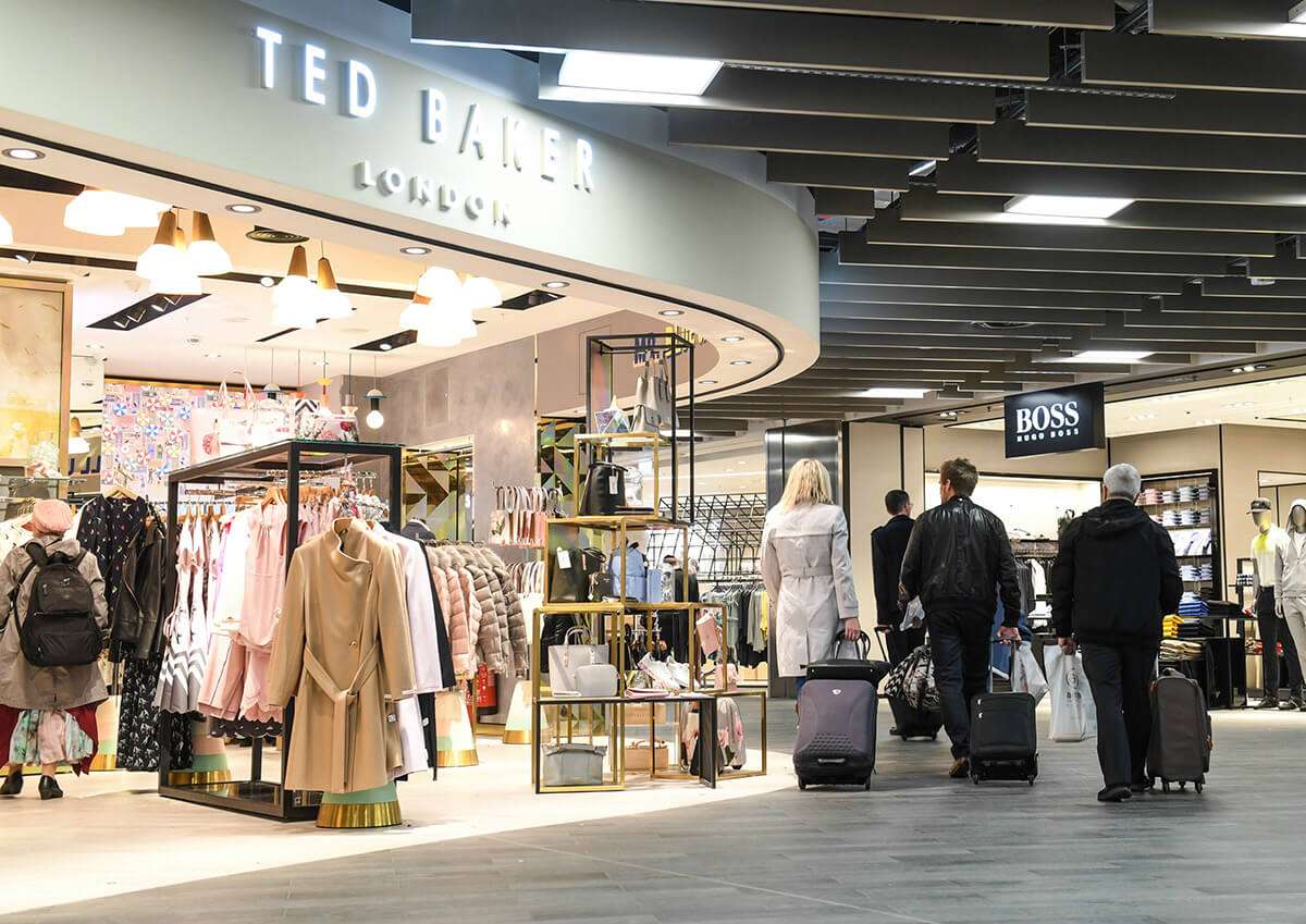 Luton Airport shops including Ted Baker and Boss, travelers with luggage browsing retail displays.